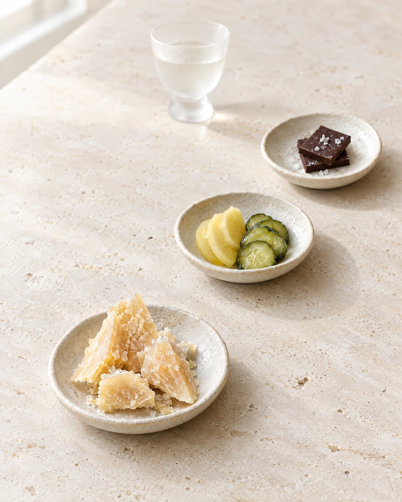 Three small ceramic dishes on pale travertine: shards of aged Parmesan, slices of pickled daikon and cucumber, and two squares of dark chocolate with sea salt. A small frosted glass of clear spirit stands beyond them.